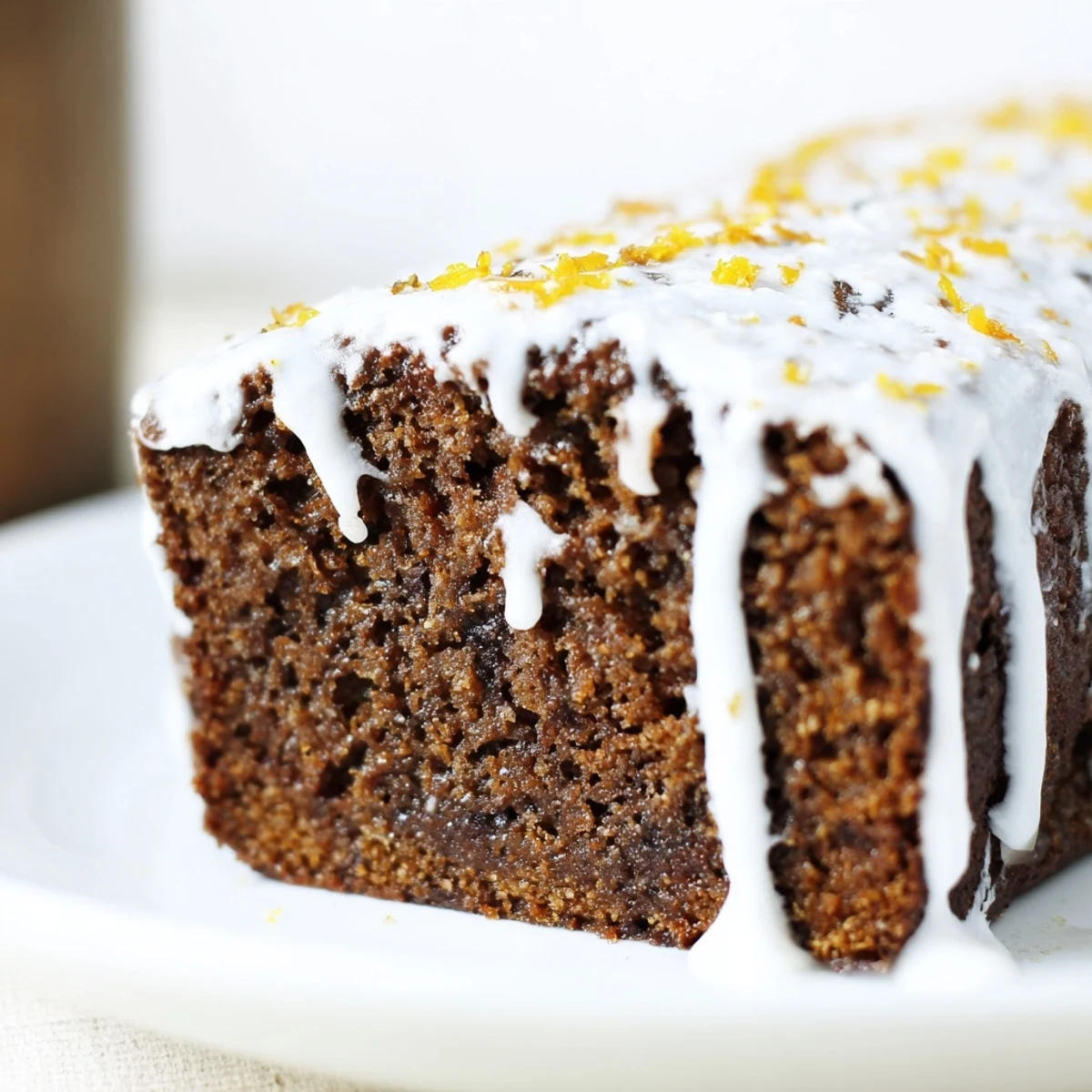 Freshly baked gingerbread loaf with orange icing cooling on a wooden board, garnished with orange zest and a cinnamon stick.