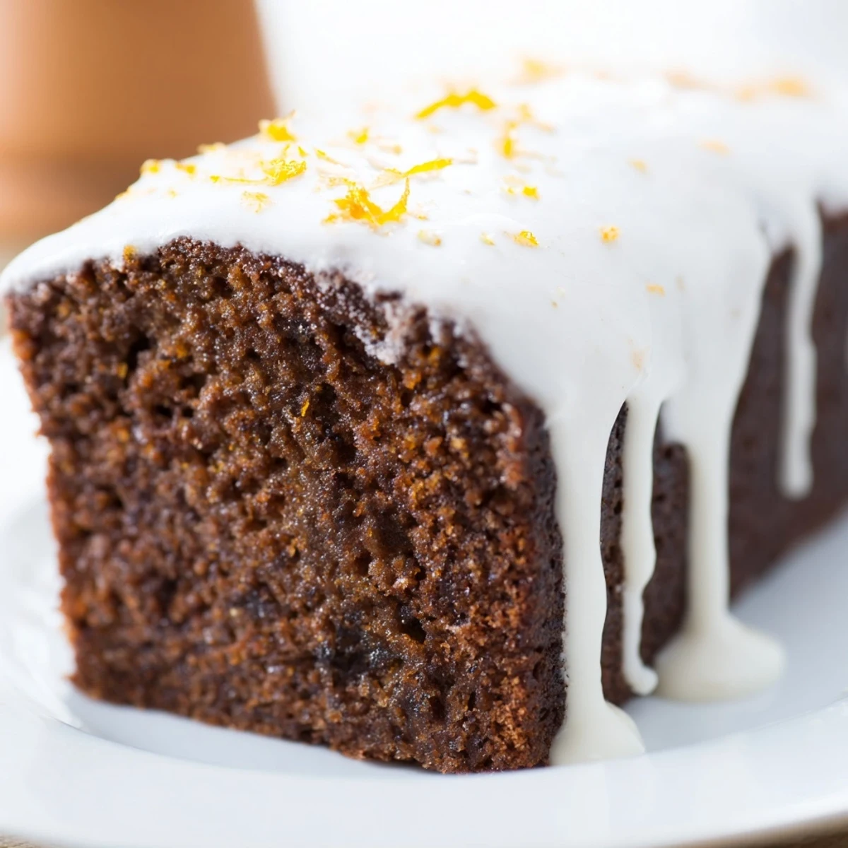 A slice of gingerbread loaf with orange icing on a white plate, next to a warm cup of tea and scattered orange peels.