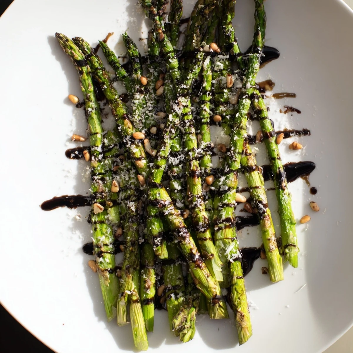 Golden roasted asparagus with balsamic glaze arranged on a white platter topped with shaved Parmesan and pine nuts.