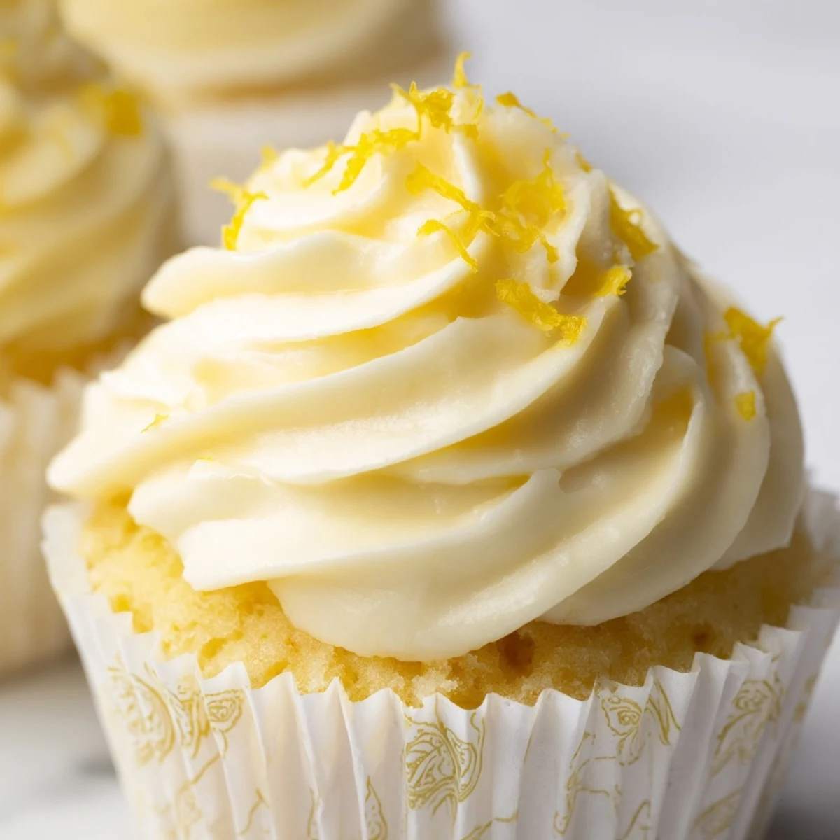 Twelve golden Limoncello Cupcakes topped with creamy frosting are arranged on a white plate beside fresh lemon slices and zest.
