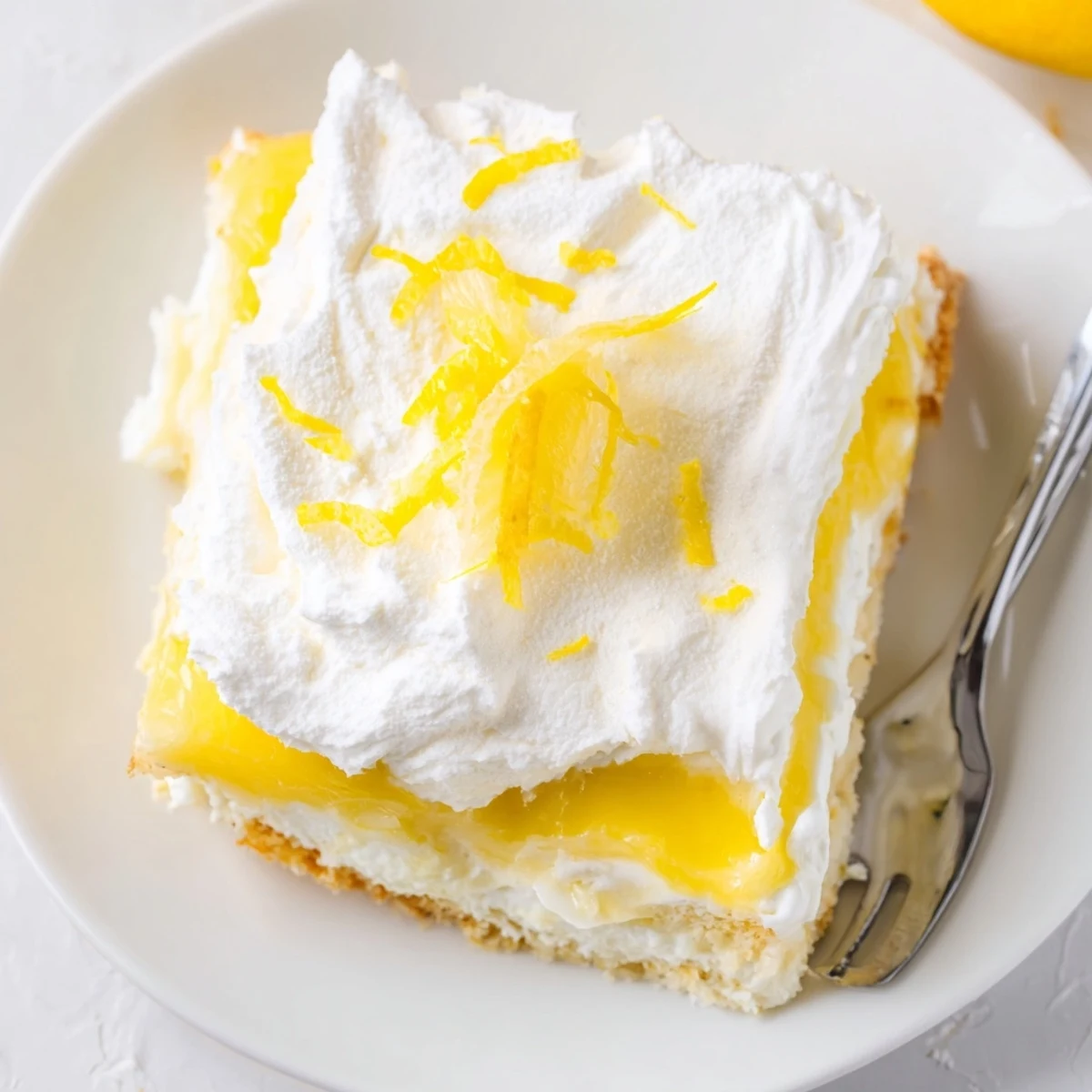 Tri-layer Lemon Lush Cake plated on white dish featuring zesty lemon pudding and creamy cheese filling with zest garnish