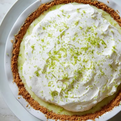 Key Lime Pie with Whipped Cream Topping is served chilled on a white plate, with a slice cut to reveal the tangy filling and buttery crust, ready for a summer dessert.  